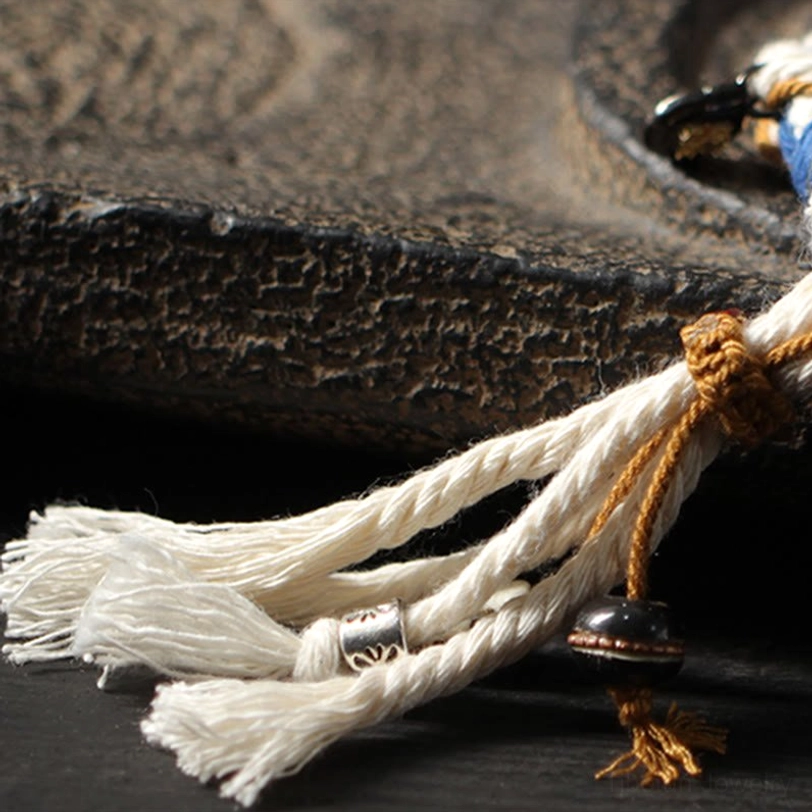 Close-up detail of Zakiram bracelet showing brown wrapping knot, silver metal charm, and fluffy cotton tassels