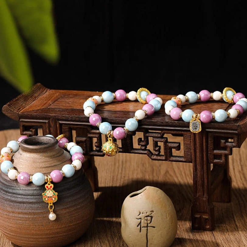 Zakiram Goddess of Wealth bracelet with enamel rabbit charm and liuli glass beads in pastel pink and blue, overhead view on vintage wooden table