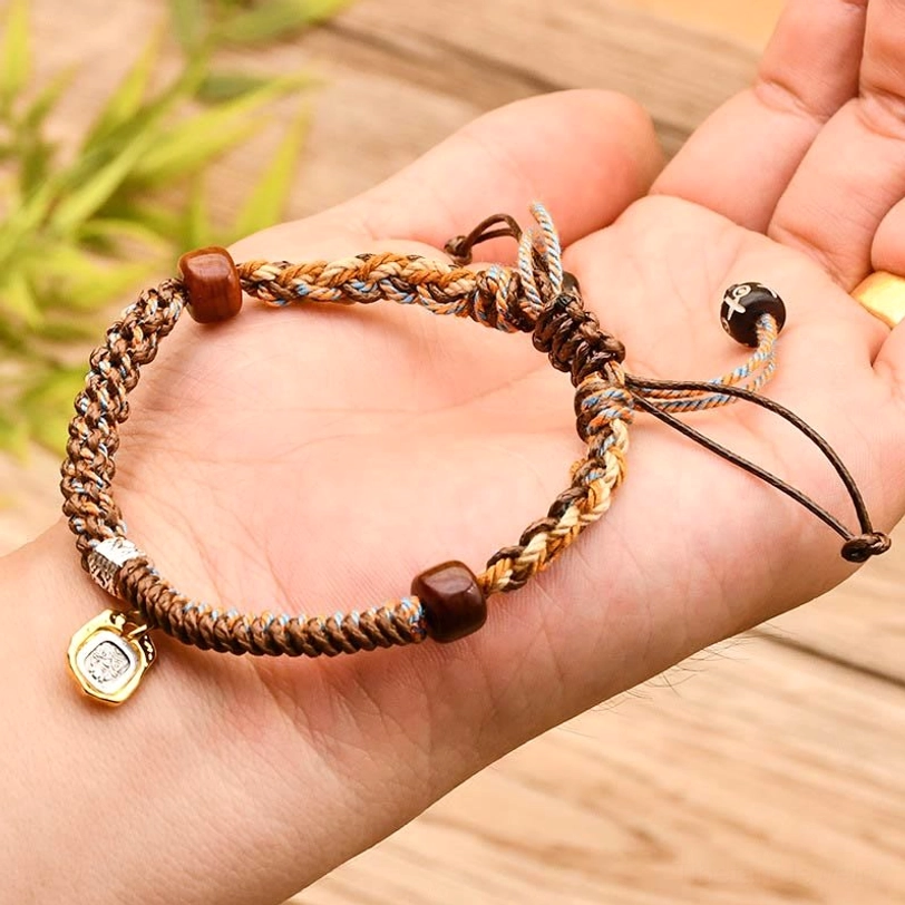 Light brown Zakiram bracelet held in palm showing detailed braided cord, wooden beads, and silver metal charm