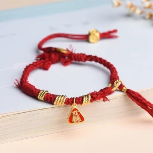 Red string Zakiram goddess protection bracelet with gold charm, displayed on book with braided cord detail
