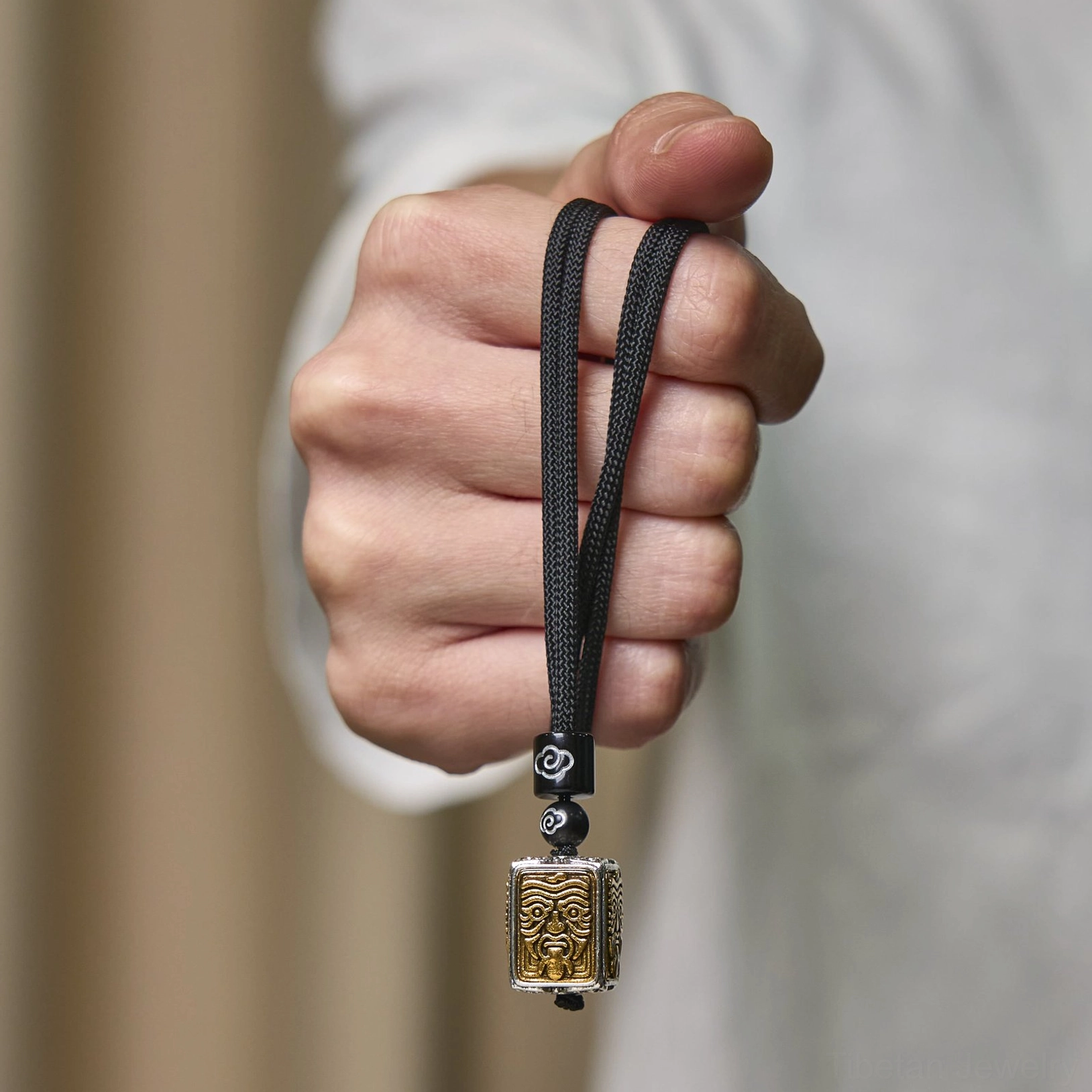 Zakiram tibetan pendant necklace held in hand, personal wearing display showing black braided rope, decorative beads, and small square bicolor deity relief pendant
